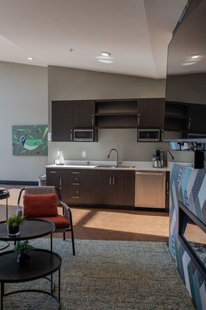 A modern kitchen with dark wood cabinets, a stainless steel sink, and a microwave. There is a red chair and a small table with a plant. - Sipped Away: King Beds, Walk in Closets (Traverse City)