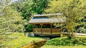 Ein Pavillon mit Dach und Holzwand, umgeben von üppigem Grün und einem kleinen Bach.
