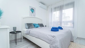 Ein modernes Schlafzimmer mit einem weißen Bett, einem kleinen Nachttisch, einer Wand-Klimaanlage und einem Fenster mit durchsichtigen Vorhängen.