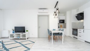 Ein modernes Wohnzimmer mit Flachbildfernseher, Esstisch und Stühlen, eine Küche mit weißen Schränken und Geräten sowie ein Teppich mit Muster.