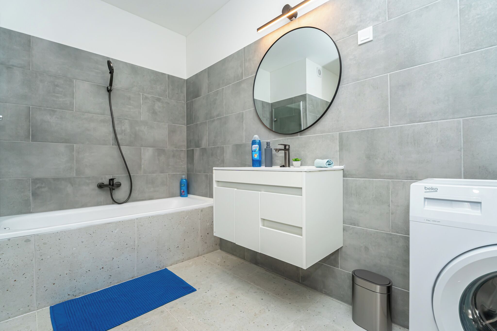 A modern bathroom with a bathtub, a washing machine, a sink with a mirror, and a trash bin.