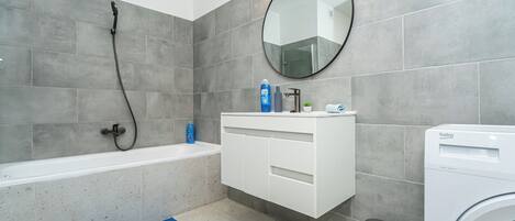 A modern bathroom with a bathtub, a washing machine, a sink with a mirror, and a trash bin.