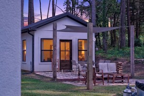A modern house with large windows, a patio with outdoor furniture, and a forested backdrop. - Stylish Farmstay on 6 Acres • Cowboy Pool • Fresh Eggs • Casita • Fire Pit (Tyler)