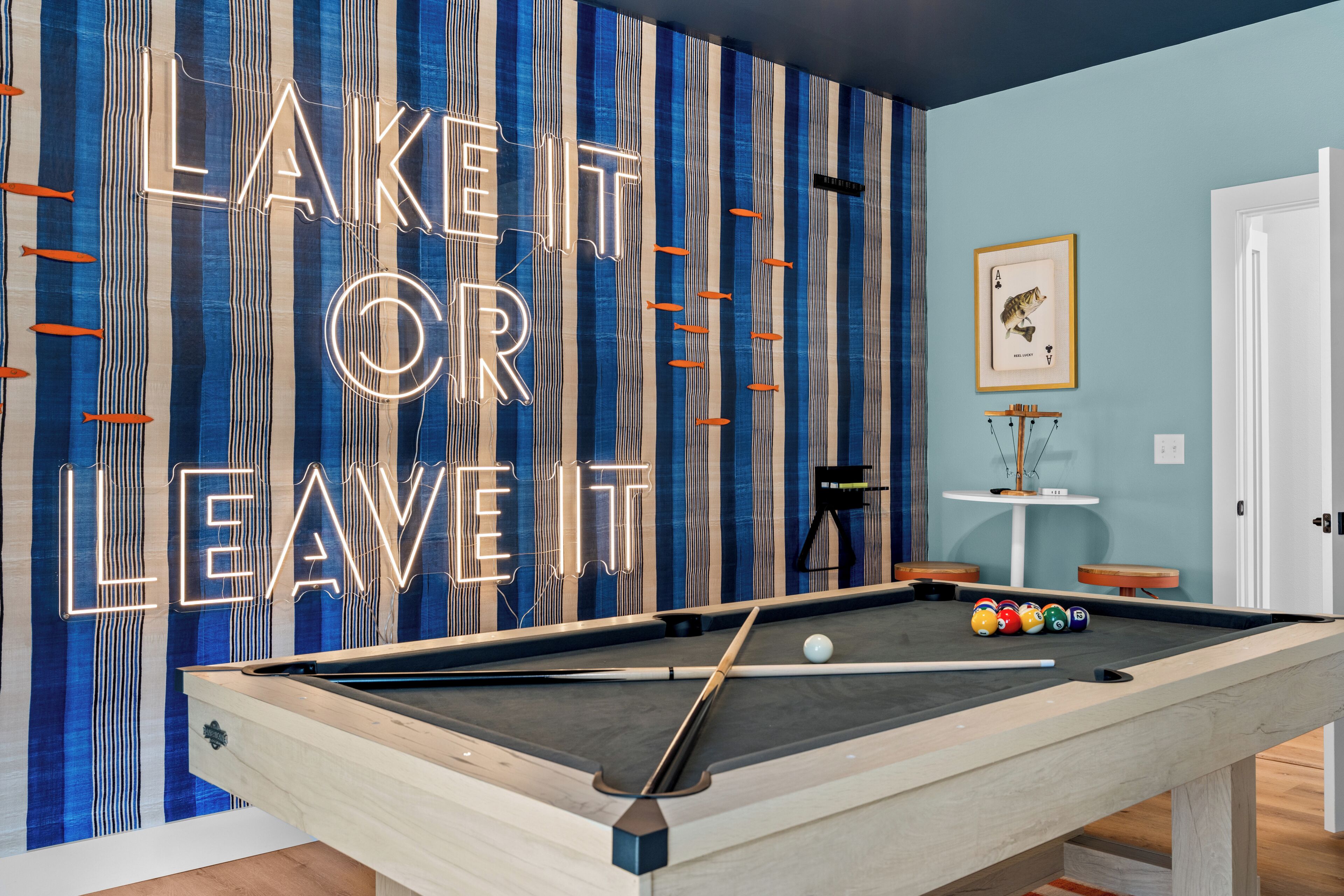 Ein Billardtisch mit einem Neon-Schild, das 'LAKE IT OR LEAVE IT' zeigt, und einer gerahmten Spielkarte an der Wand.
