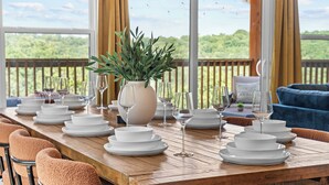 Une table à manger dressée avec de l’argenterie blanche et des verres, un vase rempli de verdure, et une vue sur un paysage verdoyant par de grandes fenêtres.