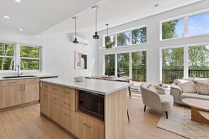 Eine moderne Küche mit einer großen Kochinsel, Holzschränken und einem eingebauten Geschirrspüler. Es gibt eine Essecke mit einer Couch und Stühlen, und große Fenster mit Blick auf Bäume.