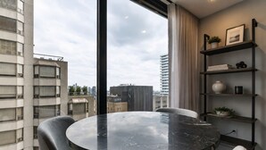 Una habitación moderna con una mesa redonda de mármol, sillas tapizadas en gris, una estantería con objetos decorativos y vistas a edificios urbanos a través de amplios ventanales.