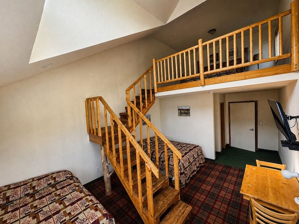 A room with a wooden staircase, two beds with patterned bedding, a plaid carpet, and a desk with a chair.