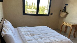 Ein ordentlich bezogenes Bett mit weißer Bettwäsche, ein kleiner Holztisch und ein Fenster mit Blick auf Bäume.