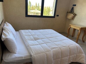 A neatly made bed with white bedding, a small wooden table, and a window with a view of trees.