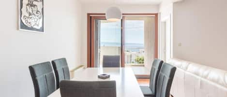 Un comedor con una mesa y cuatro sillas, una lámpara colgante, una obra de arte enmarcada en la pared y vista al mar a través de una puerta corredera.