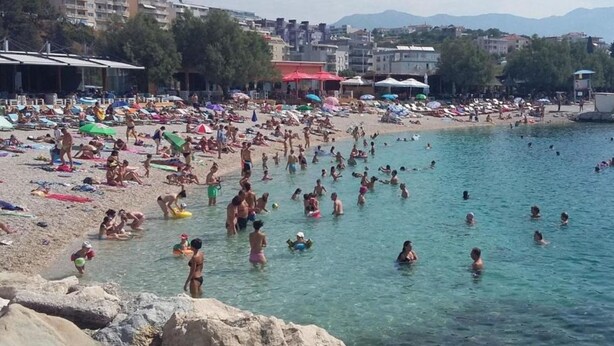 A crowded beach with people swimming and sunbathing, surrounded by buildings and mountains in the background. - Apartments Adriatic (Split)