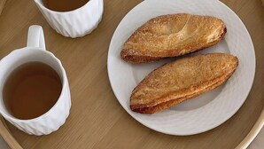 Ein Tablett mit zwei weißen Teetassen, einem Teller mit zwei Gebäckstücken und einer kleinen Topfpflanze.
