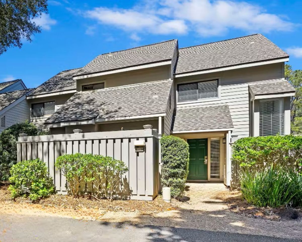 A two-story residential building with a grey roof, a green door, and a fenced front yard with shrubs.