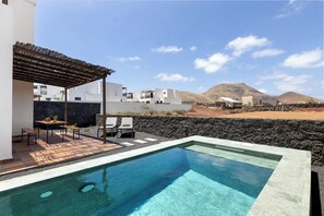 Una piscina con una pérgola, mobiliario exterior y vistas a un edificio moderno con una pared de piedra y montañas al fondo.