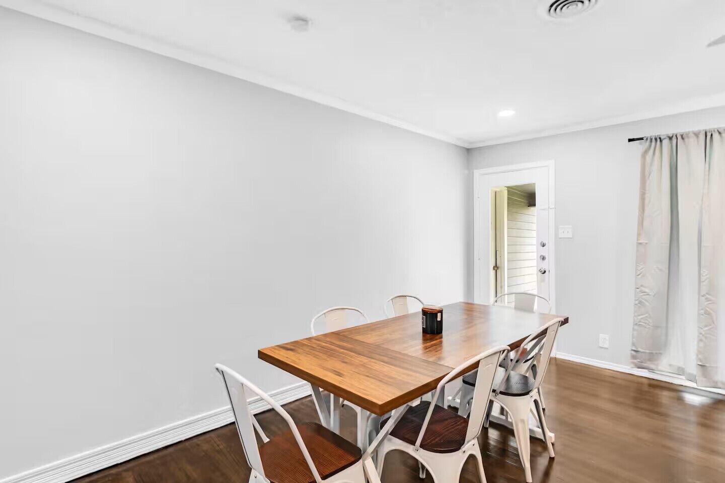 Comedor con mesa de madera y sillas blancas, pared blanca y puerta que da a otra habitación.