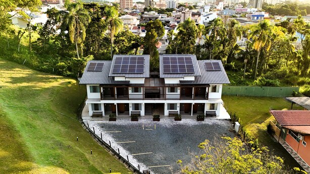 A two-story building with solar panels on the roof, surrounded by greenery and a paved area in front. - Ipê Eco Village - Apartments with Kitchen, 5 min. from Beto Carrero World (Penha)