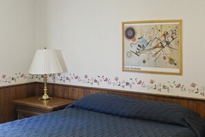 A bedroom with a bed, a nightstand with a lamp, a floral-patterned wall, and a framed abstract painting.
