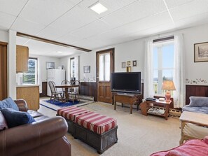 A living room with a TV, sofa, ottoman, dining table, chairs, and a kitchen area visible through an open door.