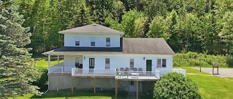 A two-story house with a covered porch and a small deck, surrounded by lush greenery.