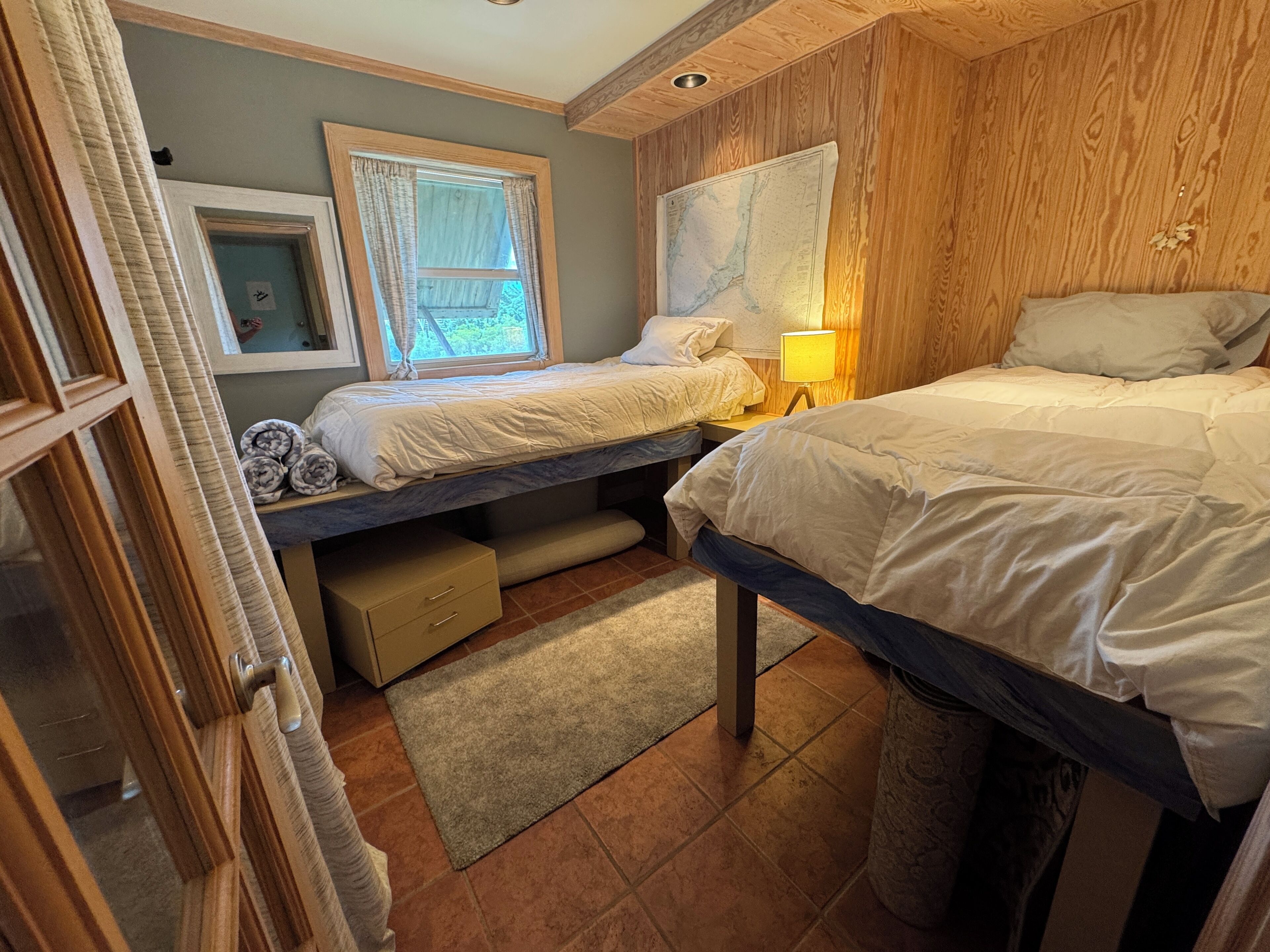 A bedroom with two beds, a window with curtains, a map on the wall, and a small lamp.