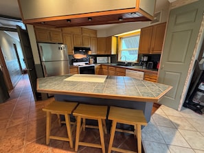 A kitchen with wooden cabinets, a stainless steel refrigerator, a microwave, a stove, and a window with a view. - Cozy, 3-Bedroom Bungalow just a short walk to beach and sound. (Rodanthe)