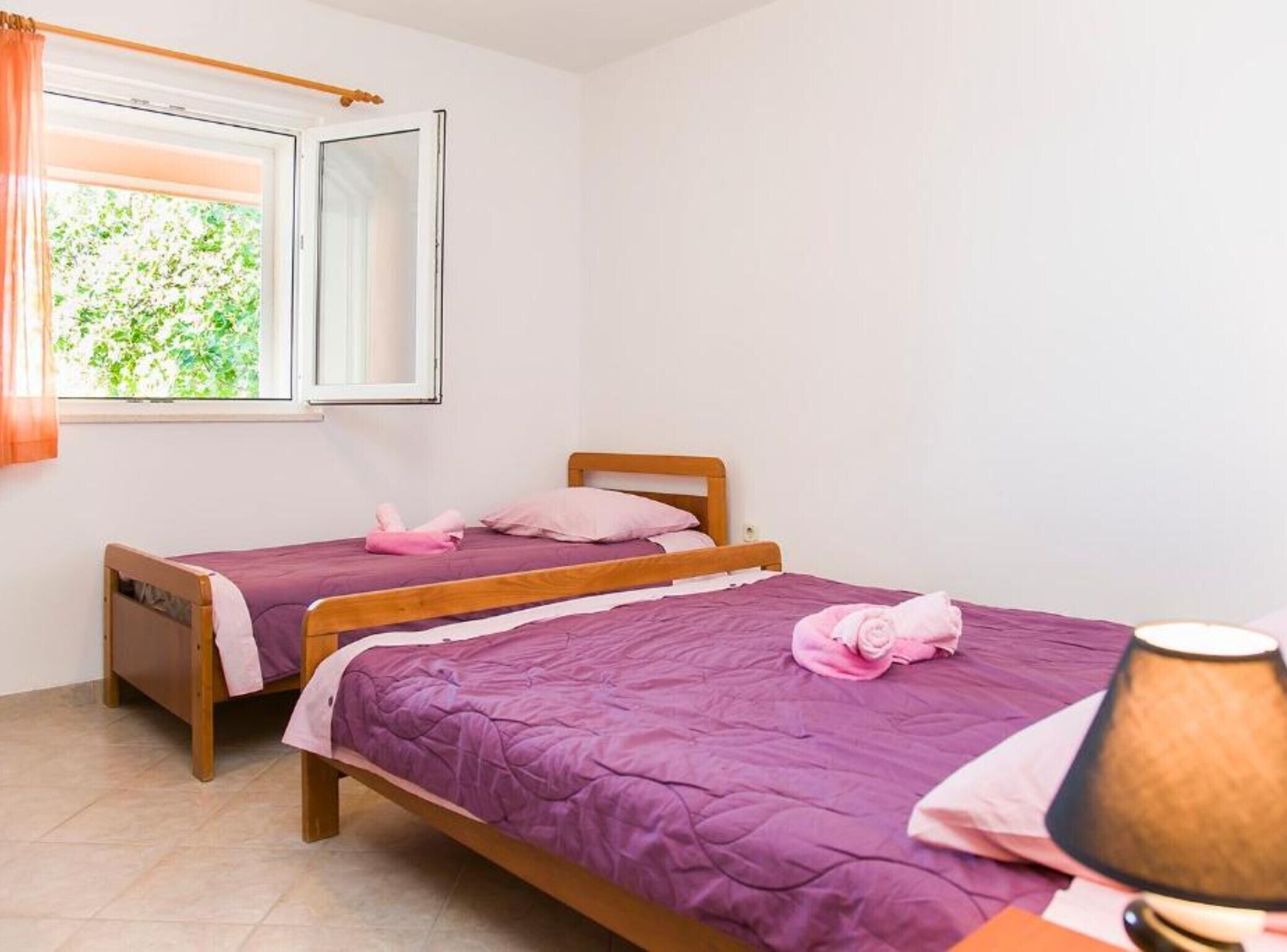 A room with two wooden beds, each with a purple bedspread and pink pillows, a window with a view of greenery, and a lamp on a side table.