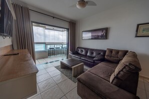 A living room with a sofa, coffee table, and a TV. There is a balcony with chairs and a view of the sea. - Apto c Varanda Vista Mar p 6 Pessoas em Cai ara (Praia Grande)