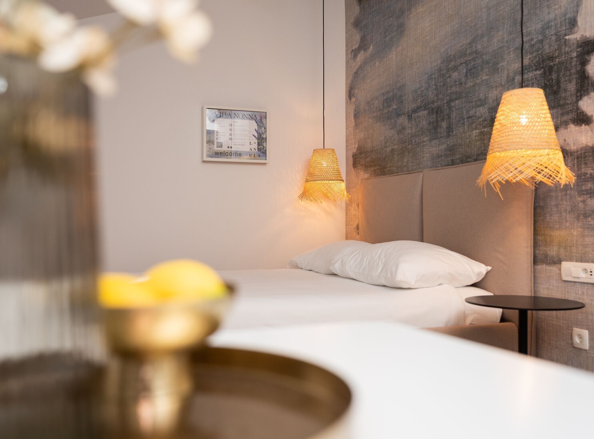 A hotel room with a bed, a small round table, a wall-mounted lamp, and a framed welcome sign.