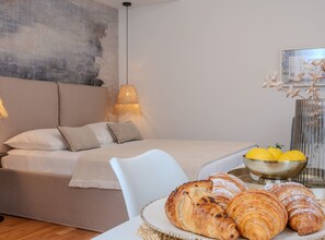A bedroom with a bed, bedside table with croissants, and a bowl of lemons. - Comfort Studio Apartment (Komiza)