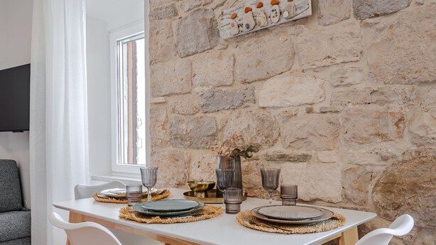 Una mesa de comedor dispuesta con platos, vasos y un florero con flores secas, frente a un muro de piedra.