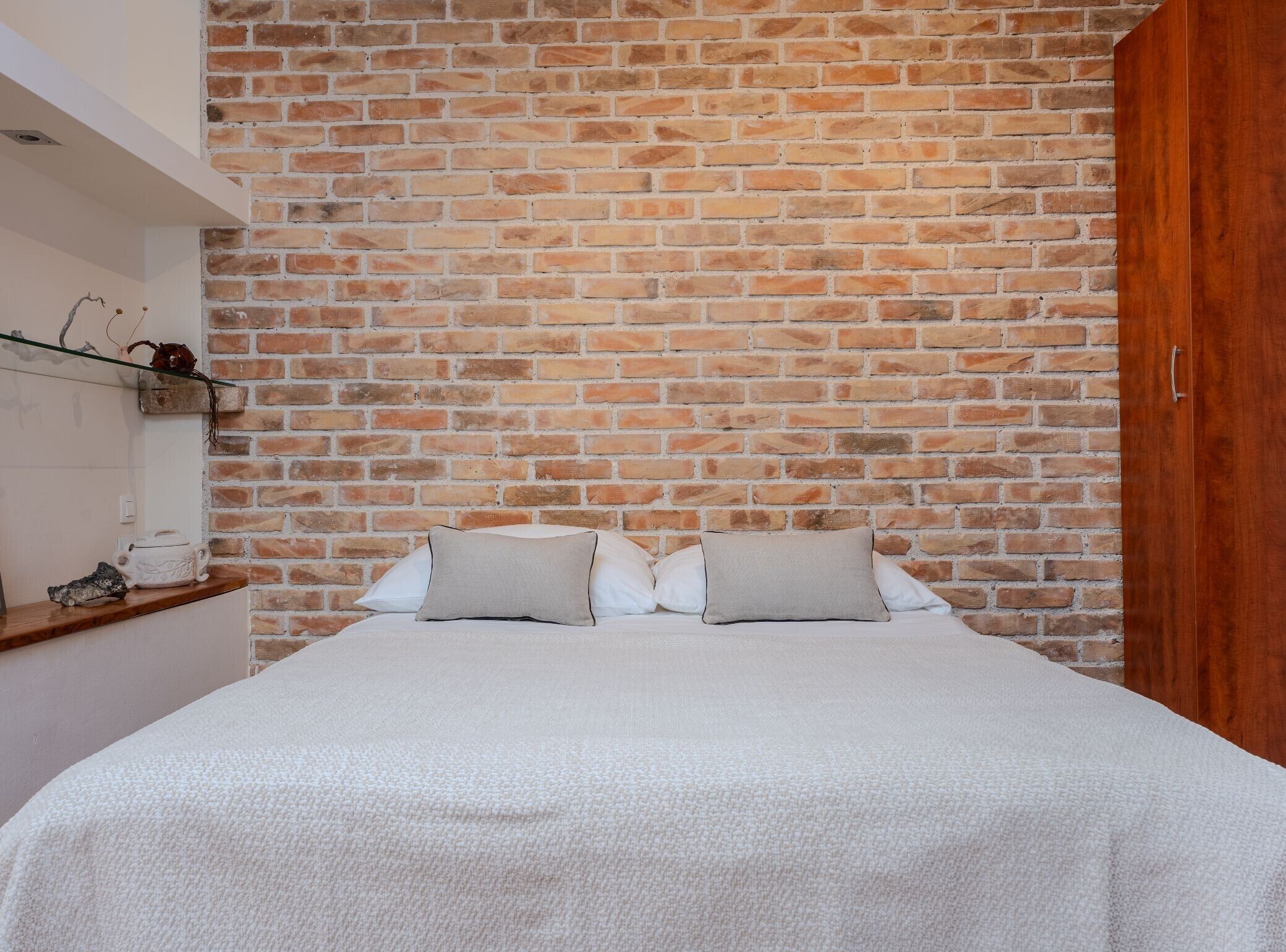 A bedroom with a brick wall, a wooden wardrobe, and a bed with white bedding.