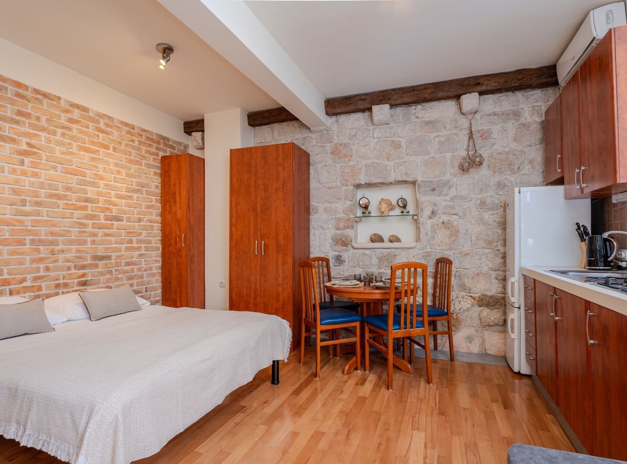 Une pièce de taille modeste comprenant un lit, une armoire en bois, une table à manger avec des chaises, un coin cuisine et un mur en pierre.