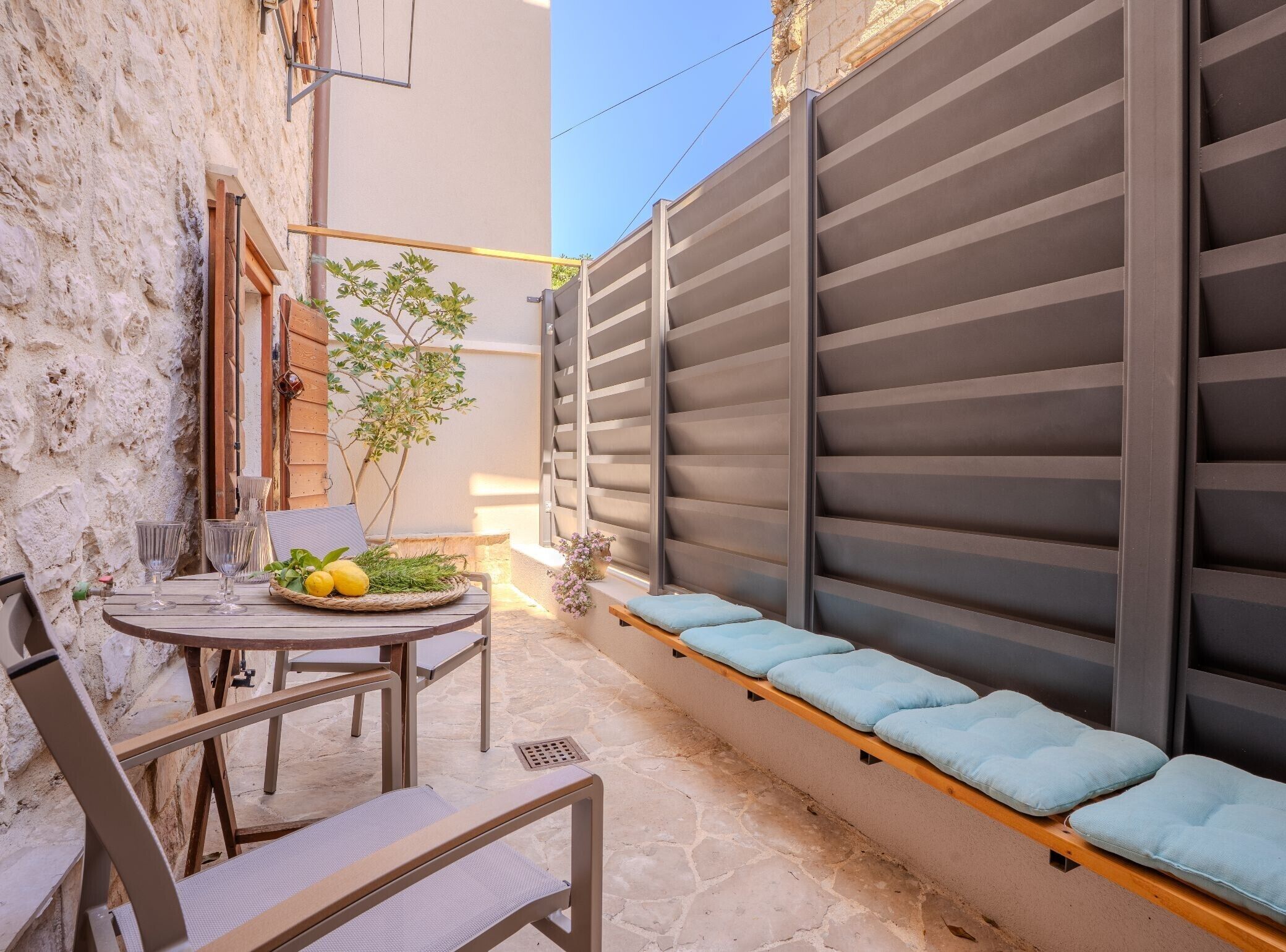 A narrow outdoor patio with a table set for two, a bench with cushions, and a potted plant.