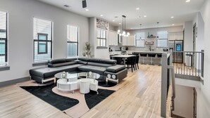 A modern living room with a sectional sofa, a dining area, a kitchen with white cabinets, and a staircase. - Brand NEW Modern Stay Near Med Center, NRG, Downtown, & more (Houston)