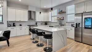 A modern kitchen with white cabinets, a marble island, black bar stools, stainless steel appliances, and a digital clock displaying 8:30.
