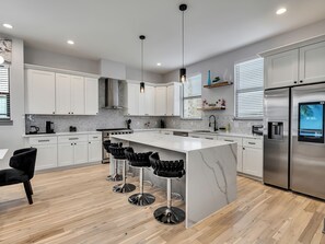 Een moderne keuken met witte kasten, een marmeren eiland, zwarte barkrukken, roestvrijstalen apparatuur en een digitale klok waarop 8:30 uur staat.