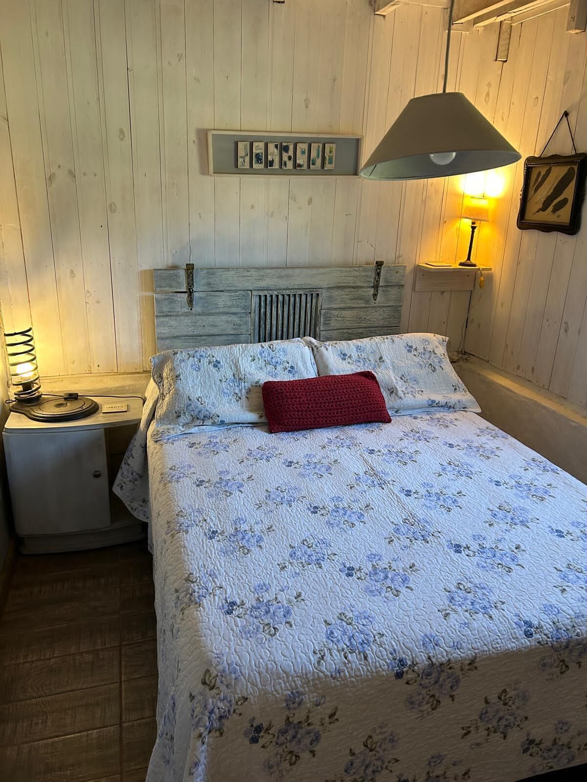 Un dormitorio con cabecera de madera, cama con colcha estampada, almohada roja, mesita de noche con lámpara y estante con marcos de fotos.