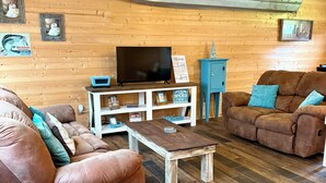 A cozy living room with a wooden ceiling, a mounted surfboard, a flat-screen TV, a coffee table, and two brown sofas with teal cushions. - Lakeside Cabin at Deer Run by Stay Current River (Van Buren)