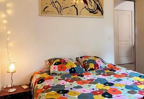 Ein Schlafzimmer mit farbenfroher floraler Bettdecke, einer Nachttischlampe und einem Wandbild.