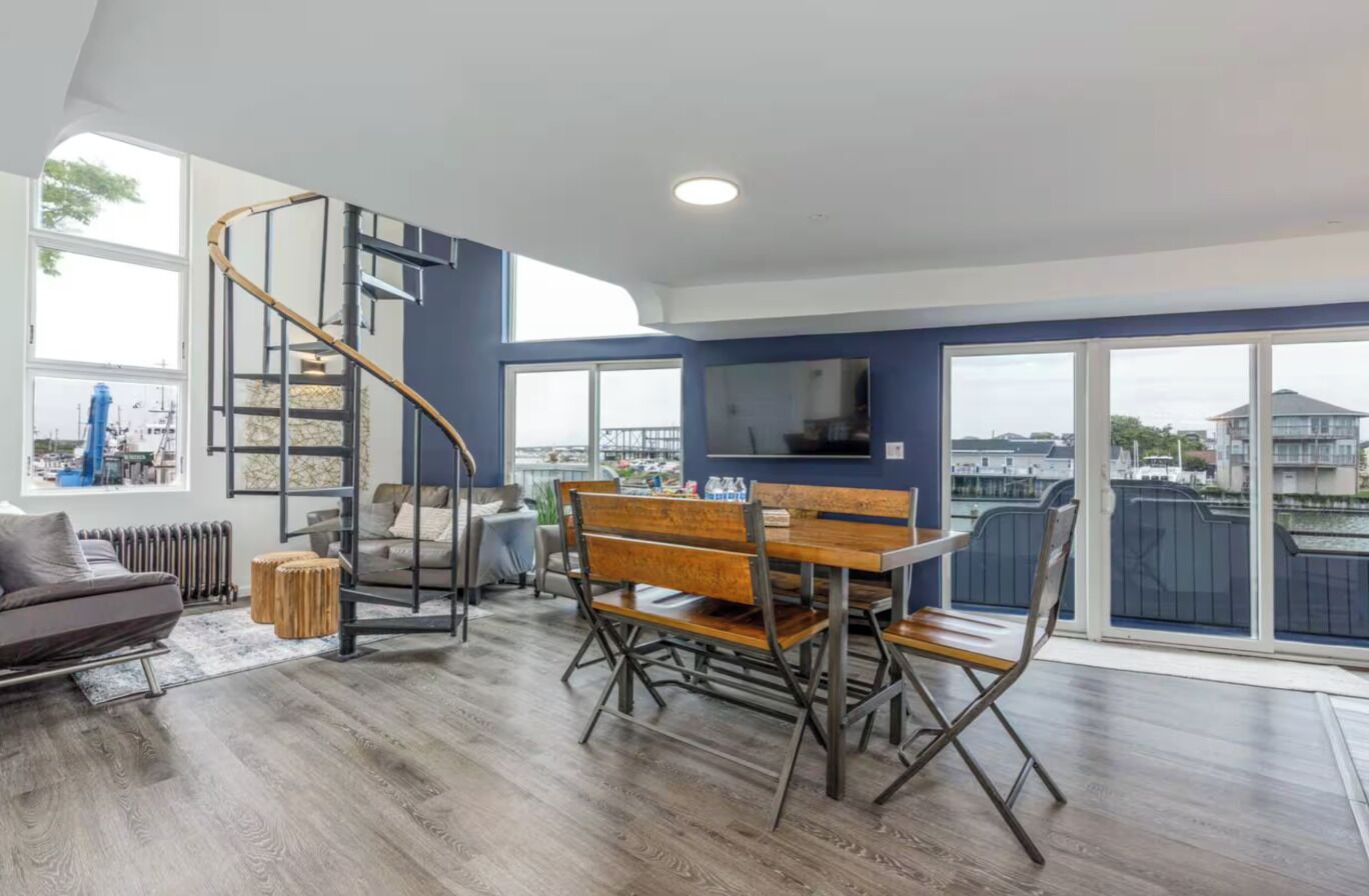A modern living room with a wooden dining table, a spiral staircase, and large windows offering a view of the city.