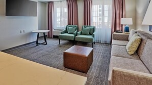 A hotel room with a flat-screen TV, two green armchairs, a sofa, a small table, a lamp, and a large ottoman. - 1BR Queen Suite w/ Hot Breakfast, Workspace & Pool (Albuquerque)