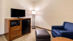 A hotel room with a flat-screen TV on a wooden cabinet, a small refrigerator, a blue sofa, an ottoman, and a floor lamp. - Comfort Suites Humble Bush Intercontinental Airport (Humble)