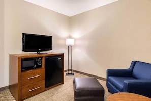 Habitación de hotel con televisor de pantalla plana sobre un mueble de madera, un refrigerador pequeño, un sofá azul, un reposapiés y una lámpara de pie.