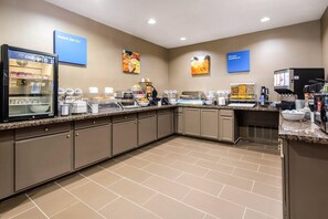 Un área de desayuno de hotel con un mostrador de autoservicio, un refrigerador y una máquina de café.