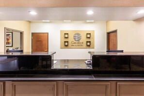 A reception area with a wooden sign that reads 'Comfort Inn & Suites.' There are two reception desks with glass tops, and a door is visible to the left. - Comfort Suites Humble Bush Intercontinental Airport (Humble)