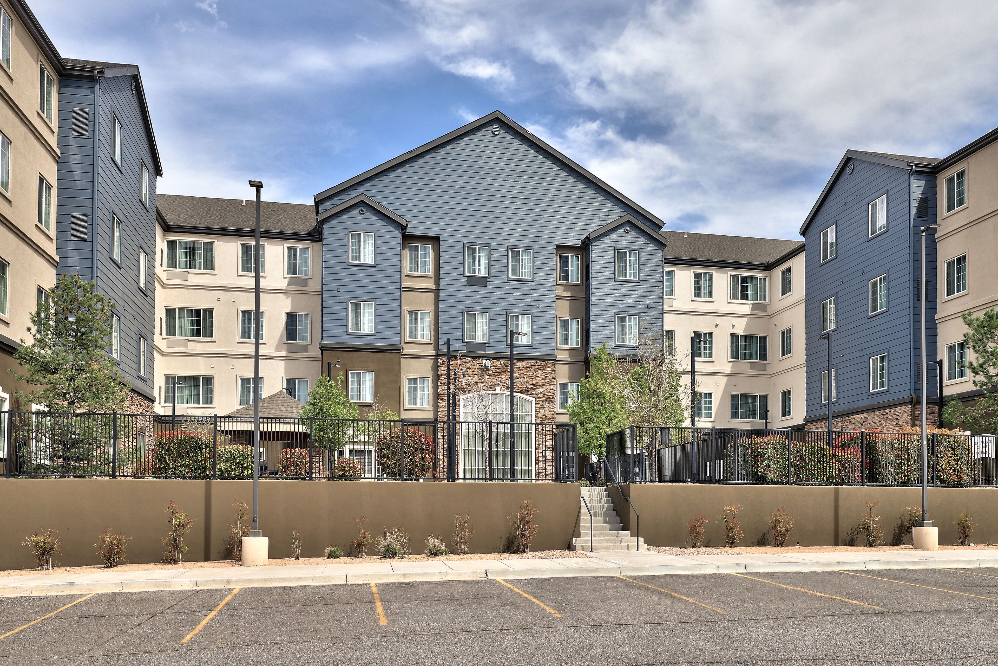 A multi-story residential building with a central entrance, a parking lot in front, and a clear sky.