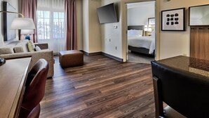 Una habitación de hotel con suelo de madera, un sofá, un escritorio, una silla, una cama, un televisor y una puerta que da a otra habitación.
