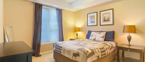 Quarto de hotel com uma cama, duas luminárias de cabeceira, uma janela com cortinas e dois quadros na parede.