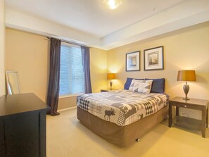 Quarto de hotel com uma cama, duas luminárias de cabeceira, uma janela com cortinas e dois quadros na parede.